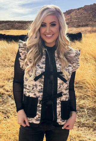 Woman wearing a patterned vest over a black top in a field with mountains in the background