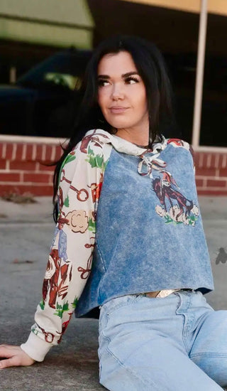 Person wearing a colorful hoodie with horse designs over a blue denim shirt, sitting on the ground.
